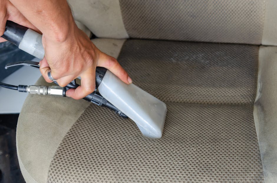 A close-up view of a person's hand holding a professional handheld upholstery cleaning machine, positioned on a beige fabric car seat with a patterned texture. The machine has a gray nozzle pressed ag